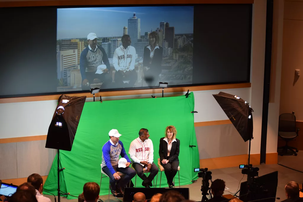 Wide shot image showcasing mcc's corporate video production service. Three people are positioned in front of a green screen, actively being recorded. The recorded image is simultaneously projected onto a large screen above them, demonstrating the real-time capabilities of mcc's setup. The image gives viewers a comprehensive look at the full video production process, emphasizing the professionalism and high-quality output of mcc's services.